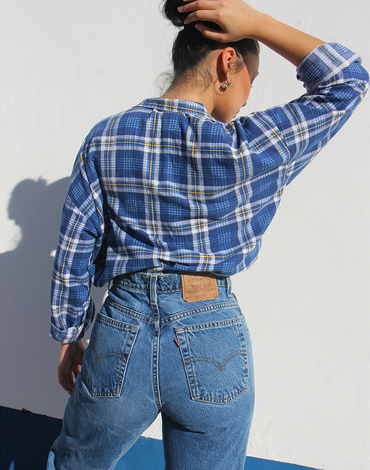 Person wearing a blue plaid shirt and jeans against a white wall