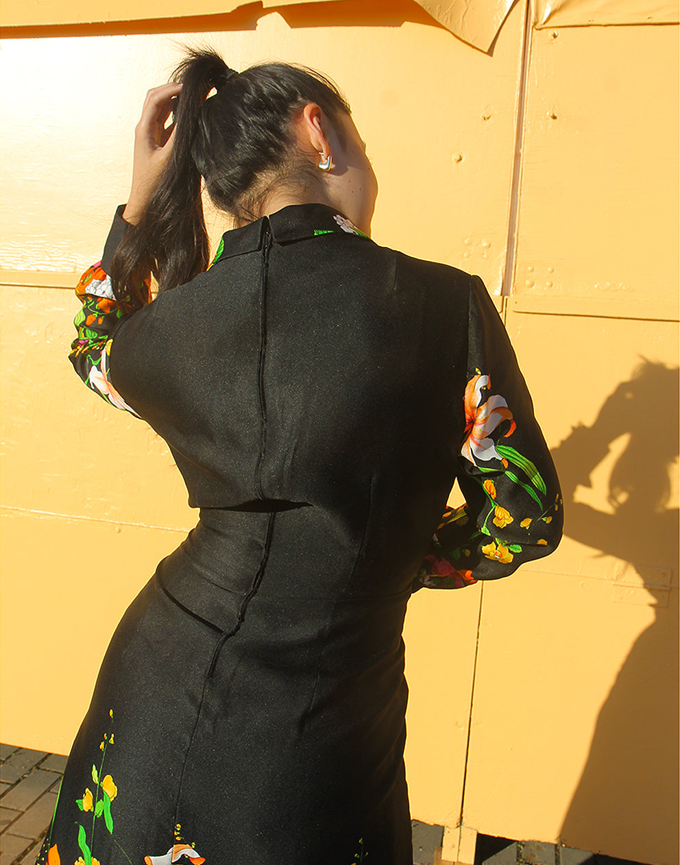 WOMAN WEARING 70S BLACK VINTAGE DRESS WITH FLORAL PRINT AGAINST A YELLOW WALL