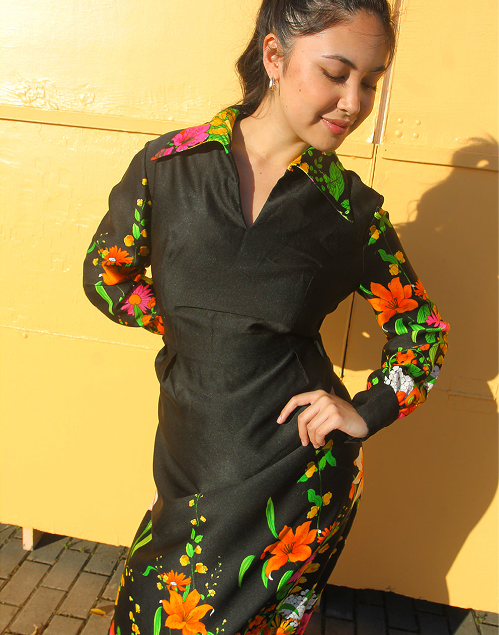 WOMAN WEARING 70S BLACK VINTAGE DRESS WITH FLORAL PRINT AGAINST A YELLOW WALL