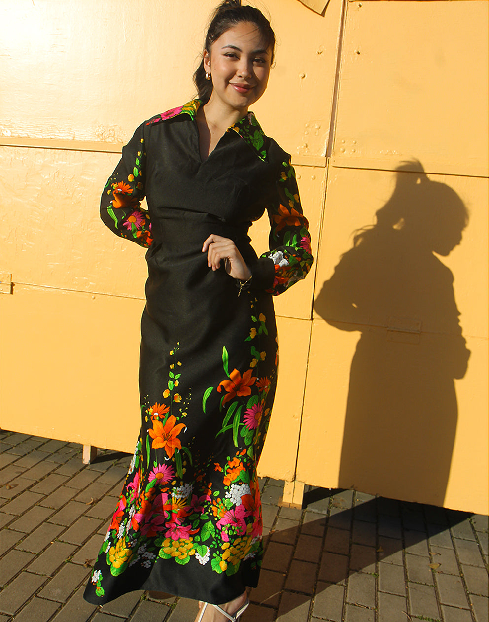 WOMAN WEARING 70S BLACK VINTAGE DRESS WITH FLORAL PRINT AGAINST A YELLOW WALL