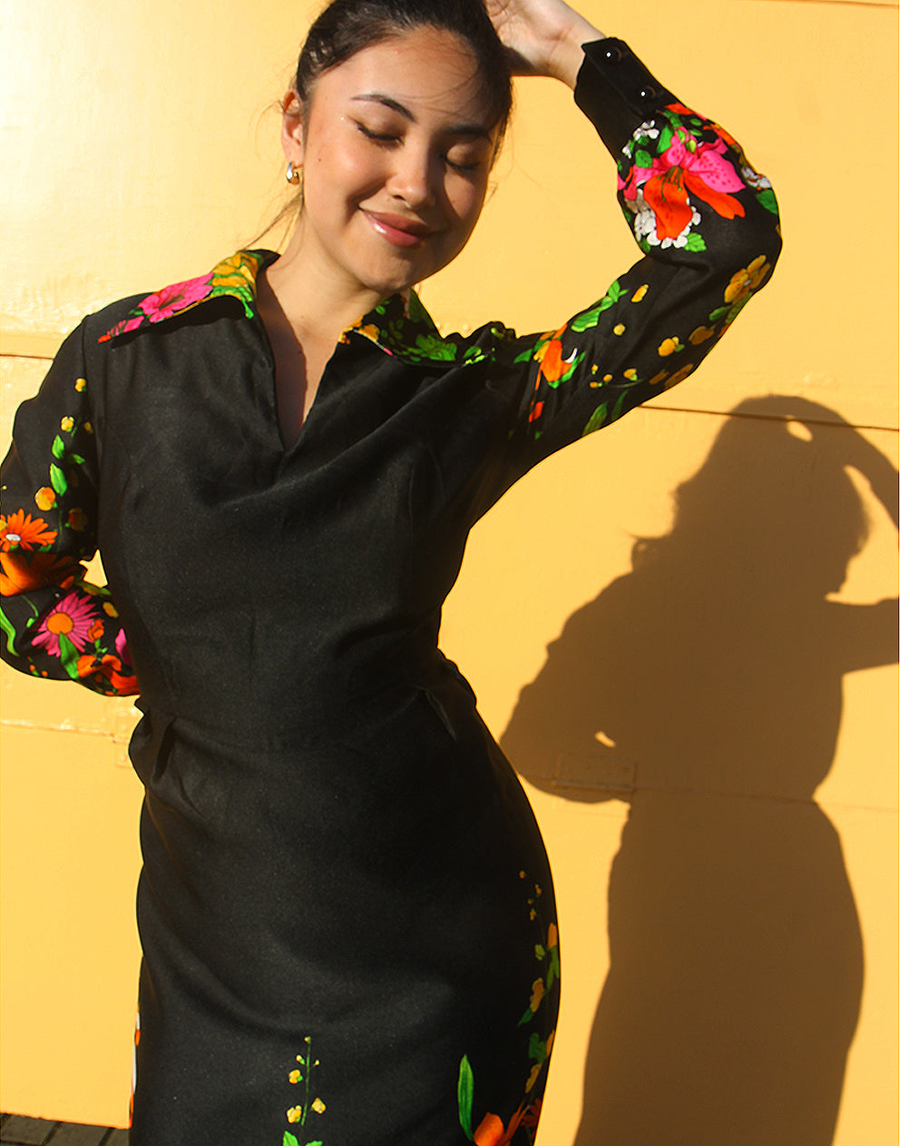 WOMAN WEARING 70S BLACK VINTAGE DRESS WITH FLORAL PRINT AGAINST A YELLOW WALL