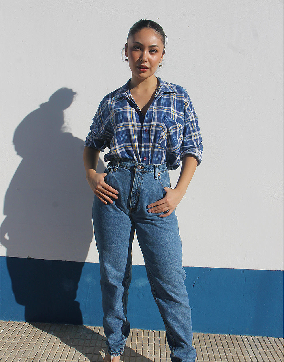 Person wearing a blue plaid shirt and jeans standing against a white wall with a blue stripe.
