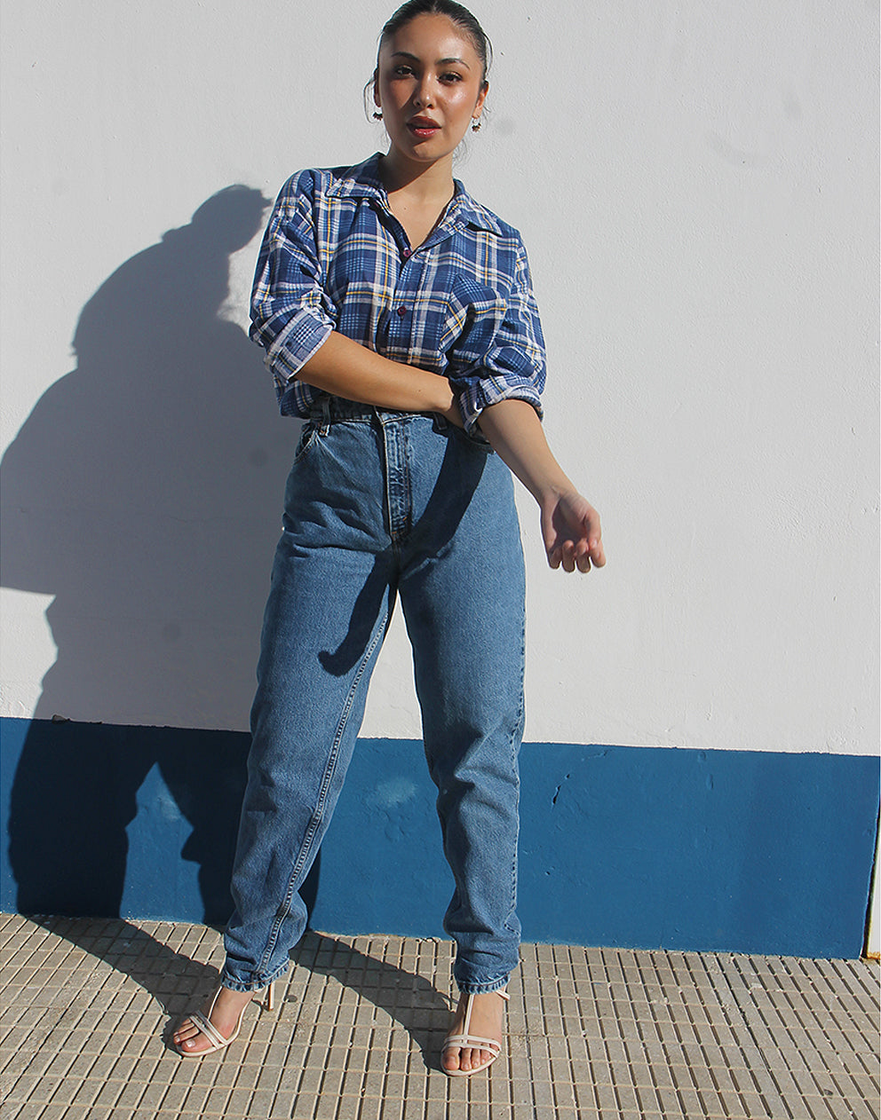 Woman wearing a blue plaid shirt and jeans standing against a white and blue wall.
