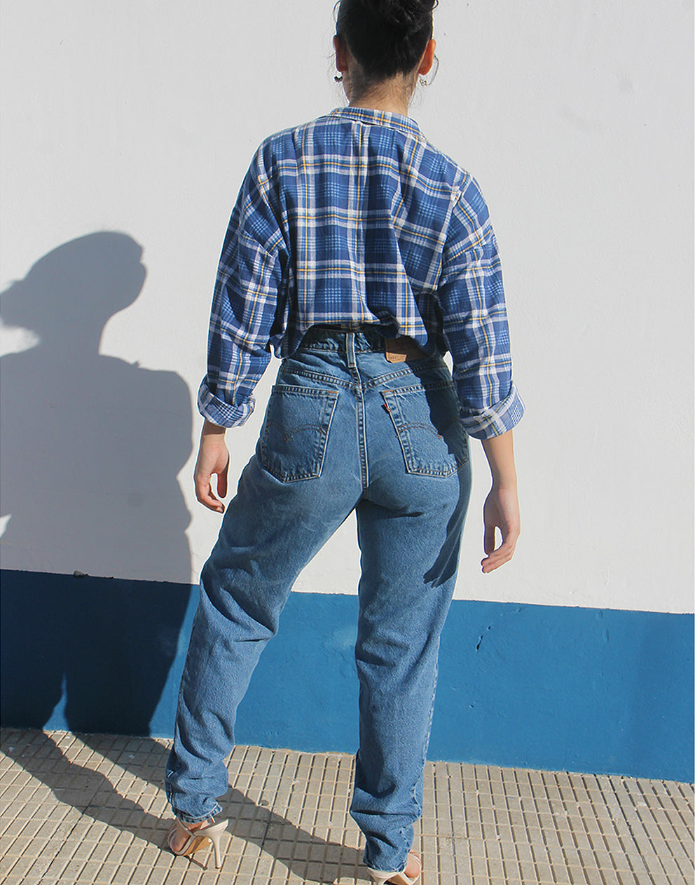 Person wearing a blue plaid shirt and jeans standing against a white wall with a blue base.