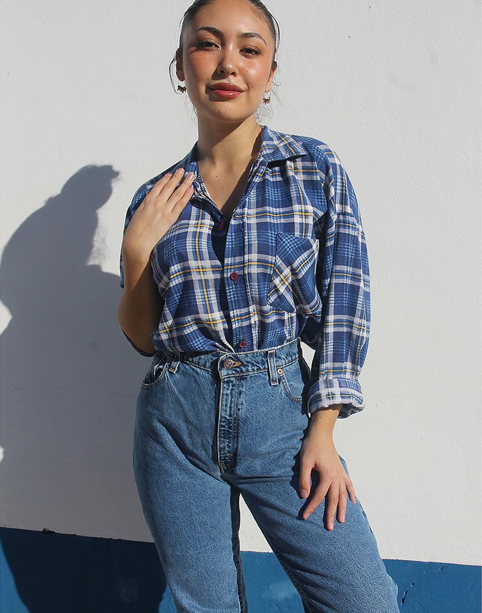 Person wearing a blue plaid shirt and jeans against a white wall