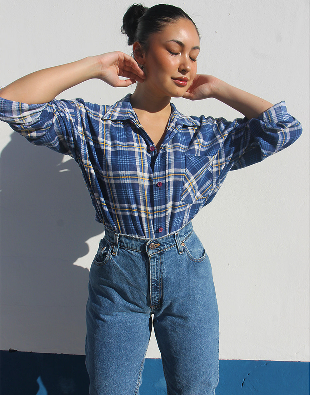 Person wearing a blue plaid shirt and jeans against a white wall