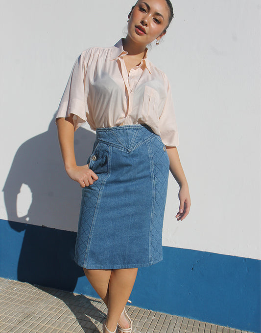 Woman wearing a light pink blouse and blue denim skirt against a white wall with a blue base.