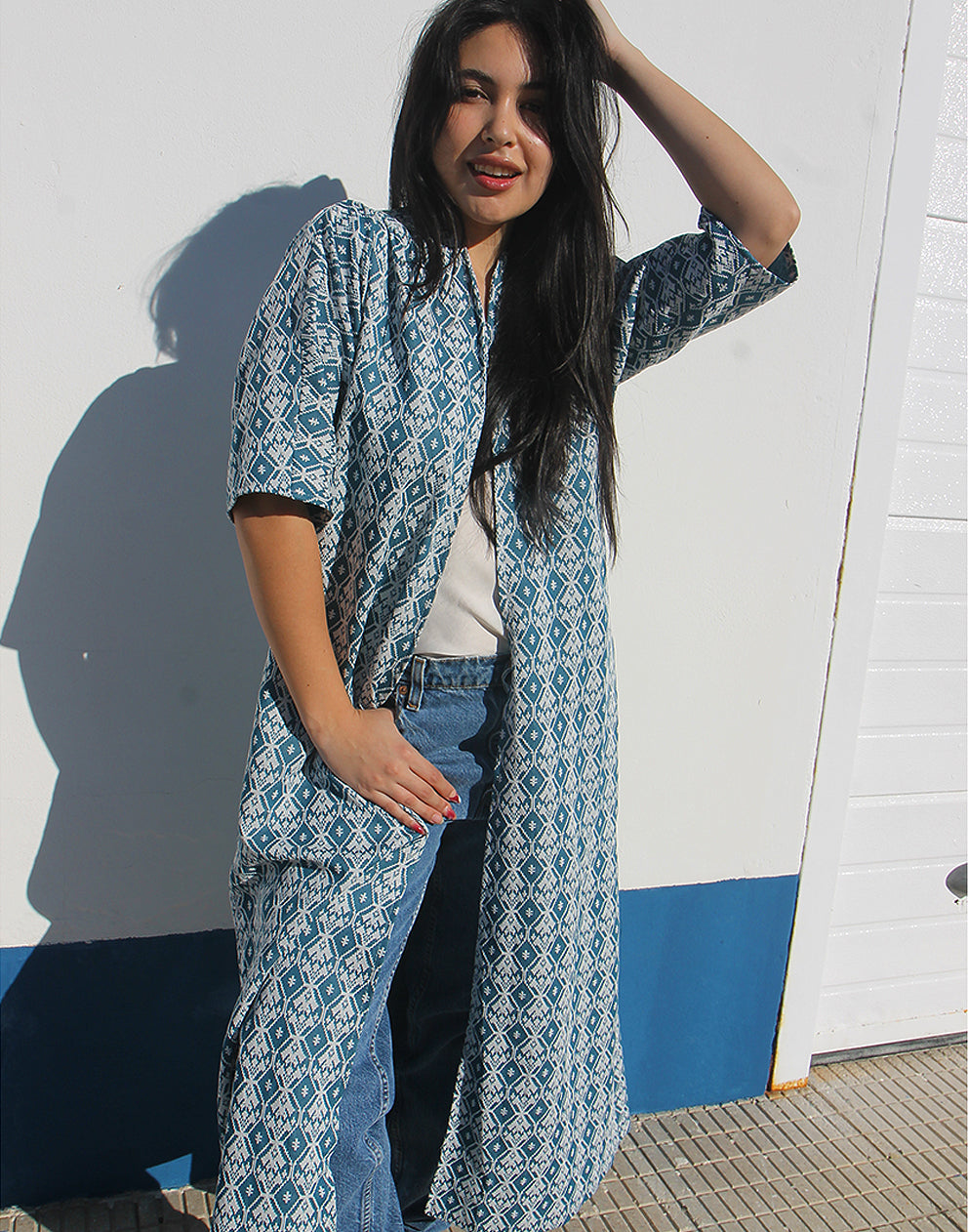 WOMAN WEARING A LONG BLUE JACKET AGAINST A WHITE WALL 