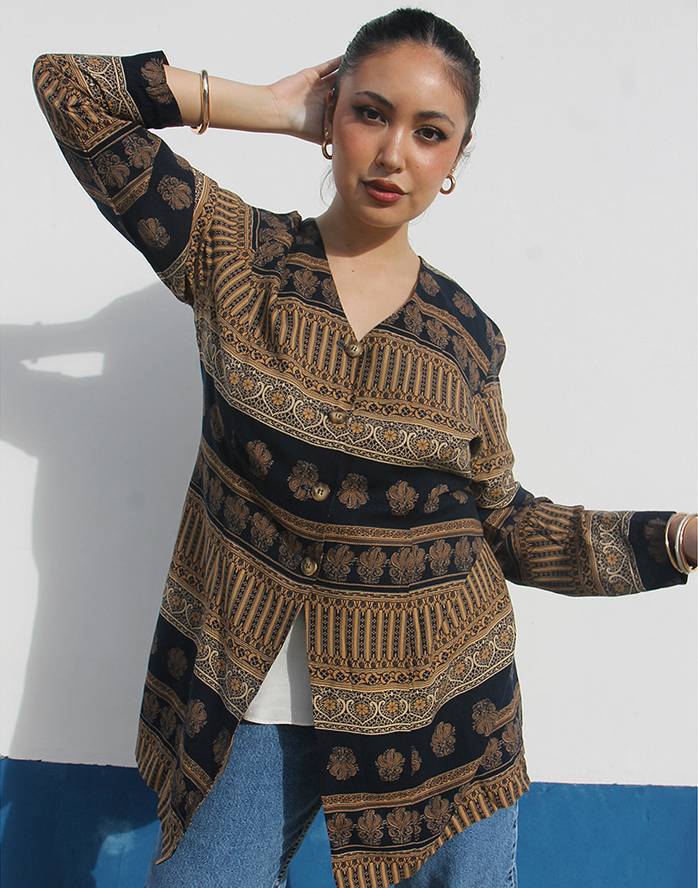 Woman wearing a brown and black printed long shirt and blue jeans against a white wall