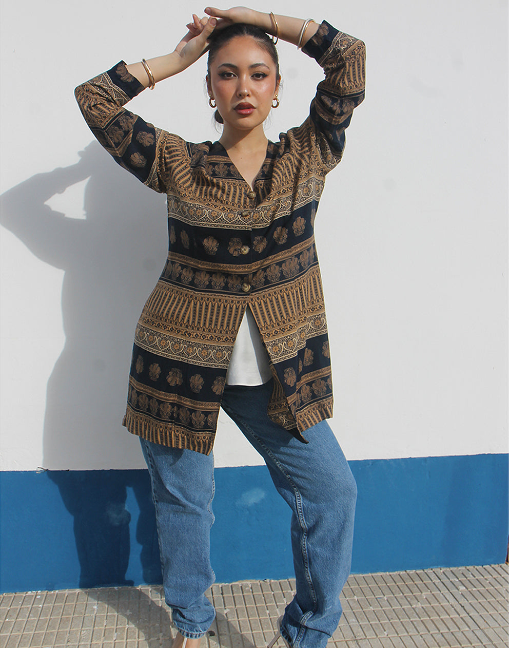 Woman wearing a brown and black printed long shirt and blue jeans against a white wall