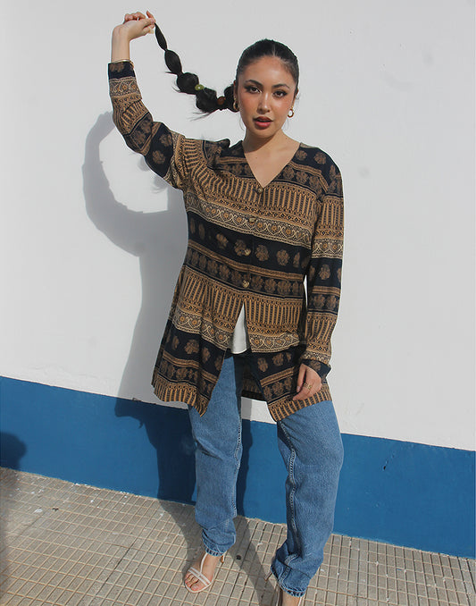 Woman wearing a brown and black printed long shirt and blue jeans against a white wall