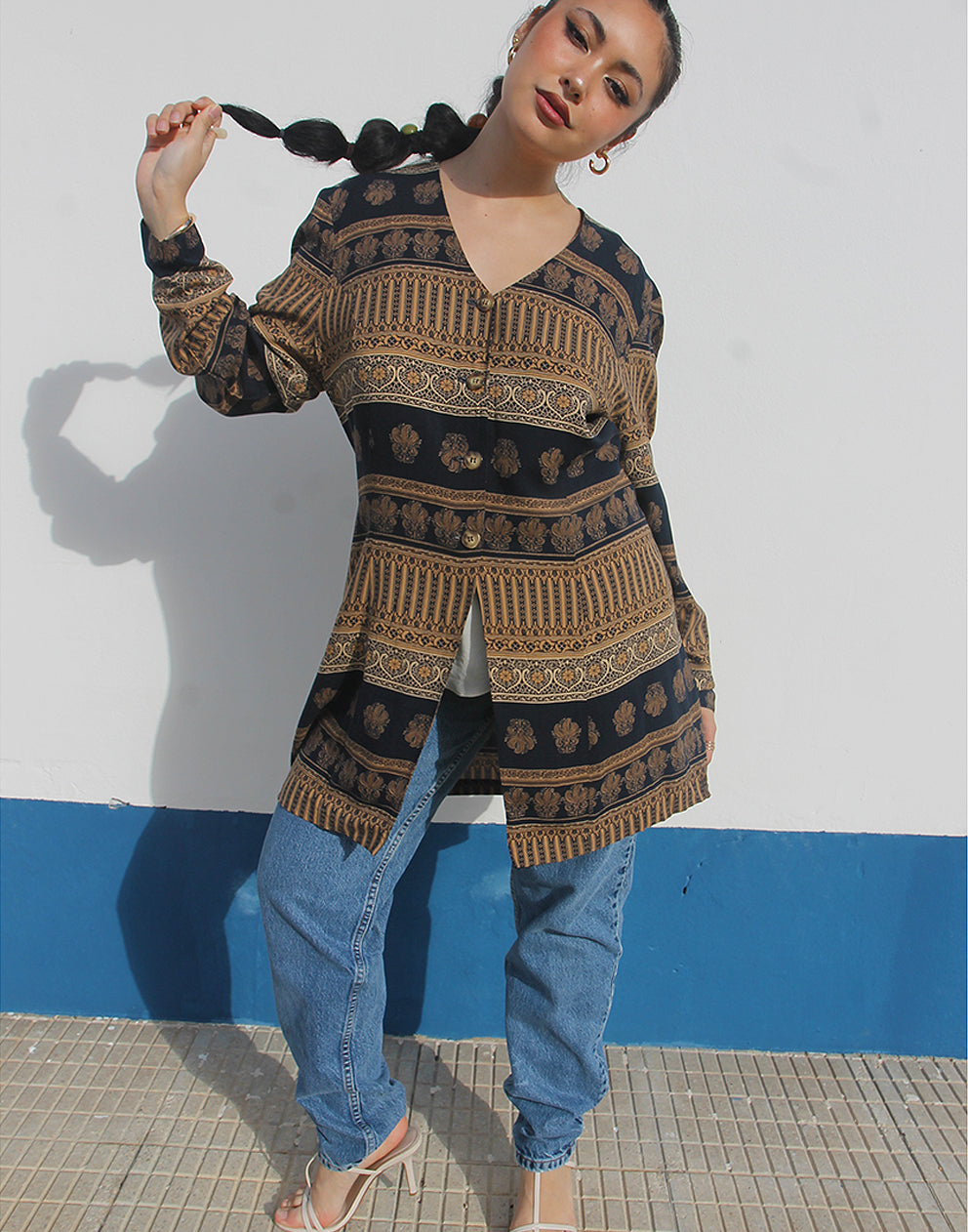 Woman wearing a brown and black printed long shirt and blue jeans against a white wall
