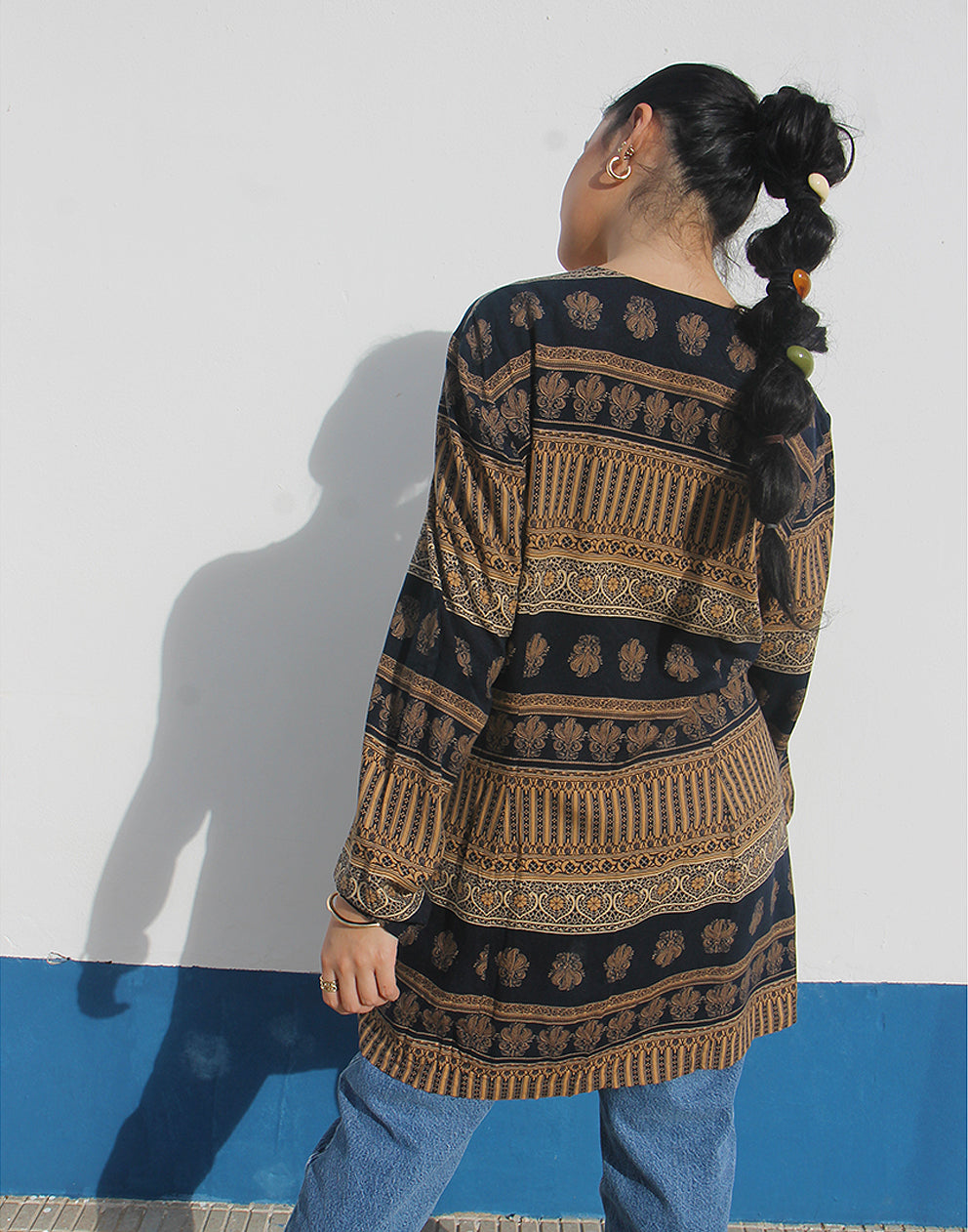 Woman wearing a brown and black printed long shirt and blue jeans against a white wall