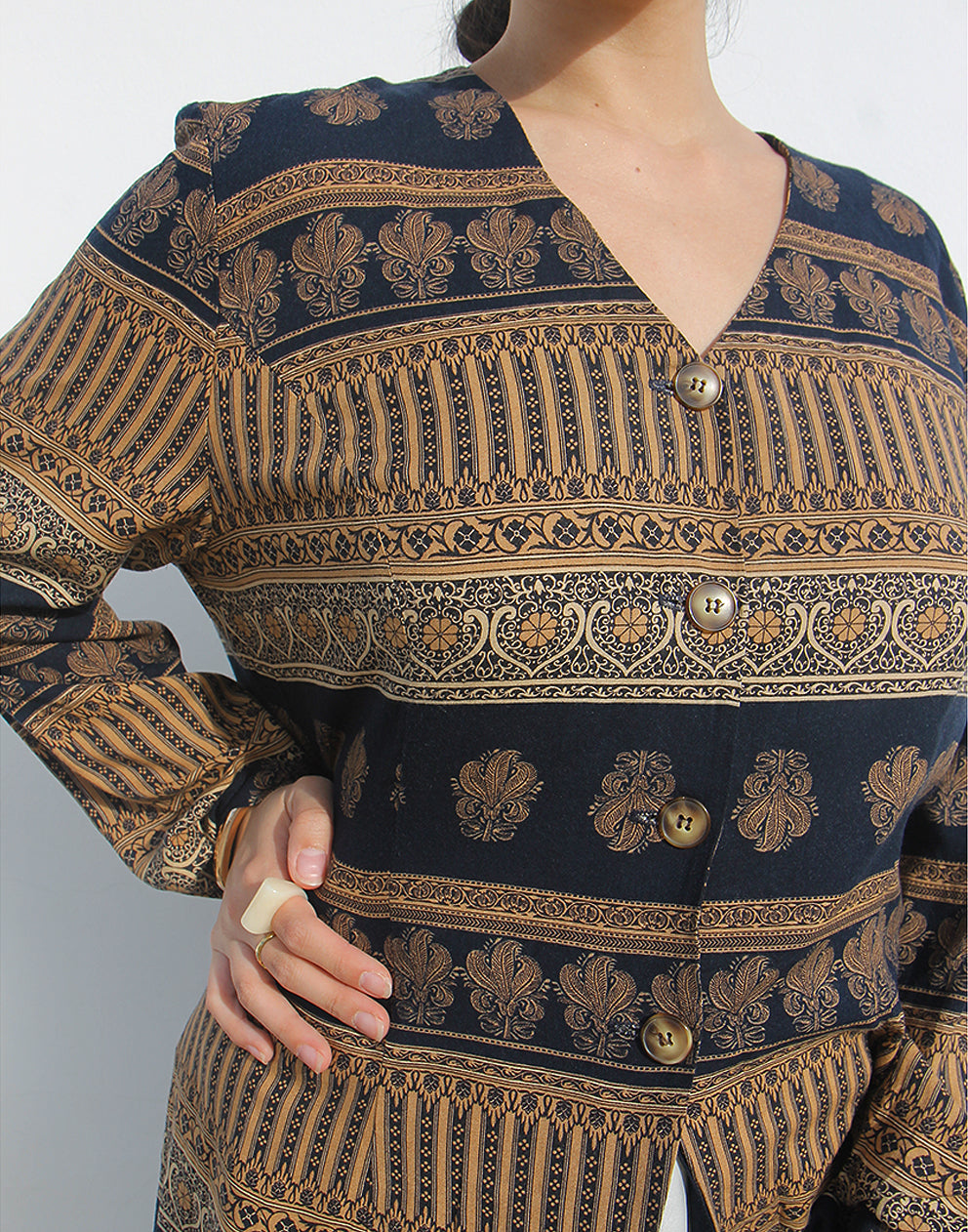 Woman wearing a brown and black printed long shirt and blue jeans against a white wall