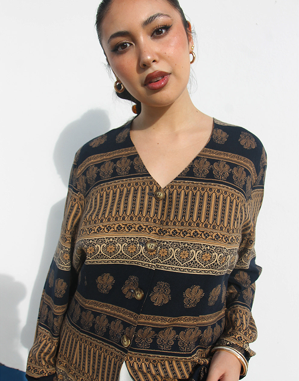 Woman wearing a brown and black printed long shirt and blue jeans against a white wall