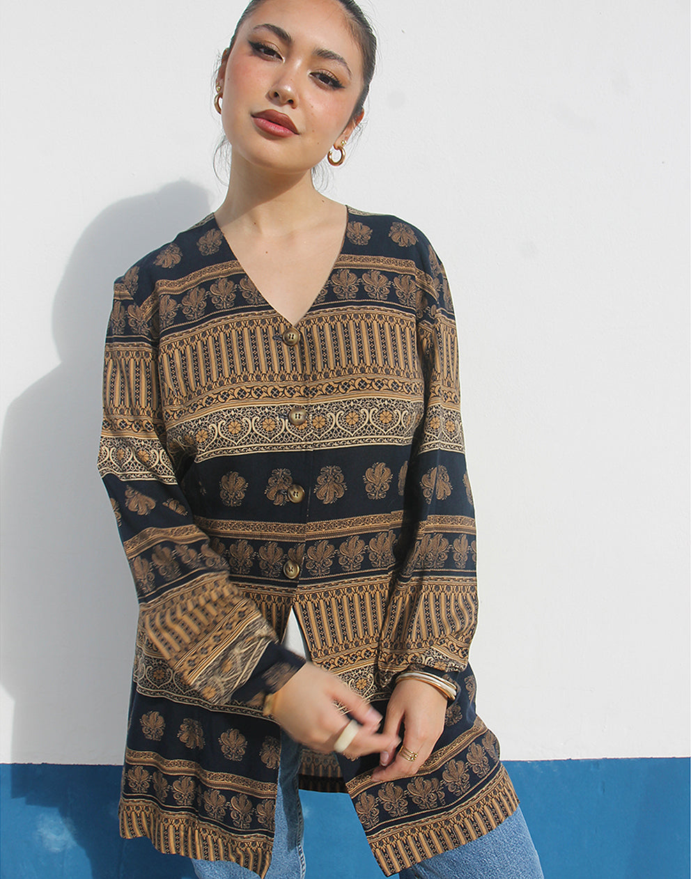 Woman wearing a brown and black printed long shirt and blue jeans against a white wall
