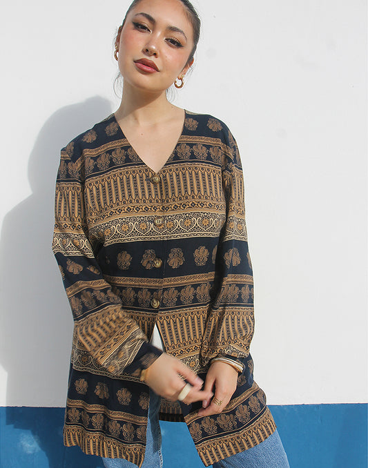 Woman wearing a brown and black printed long shirt and blue jeans against a white wall