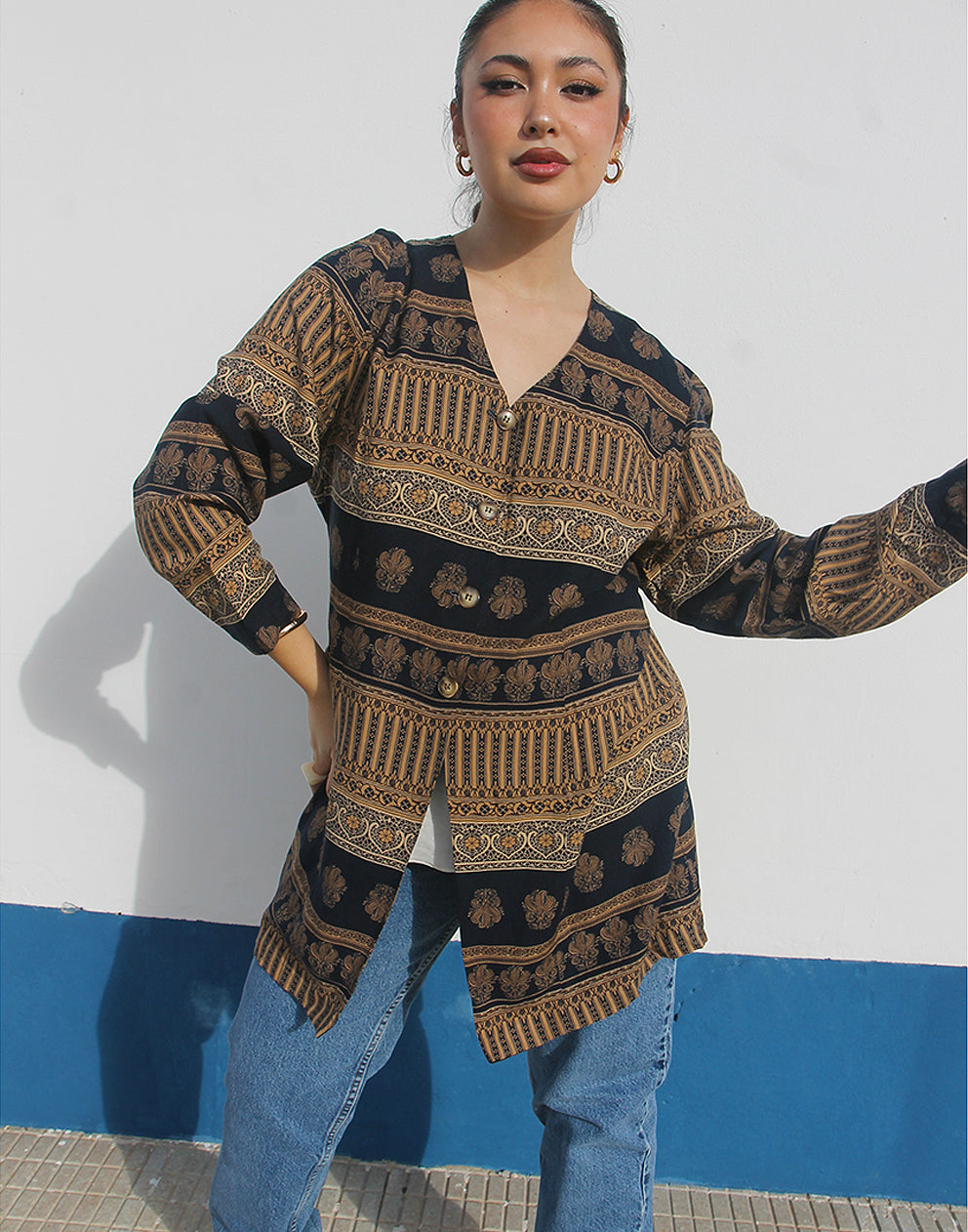 Woman wearing a brown and black printed long shirt and blue jeans against a white wall